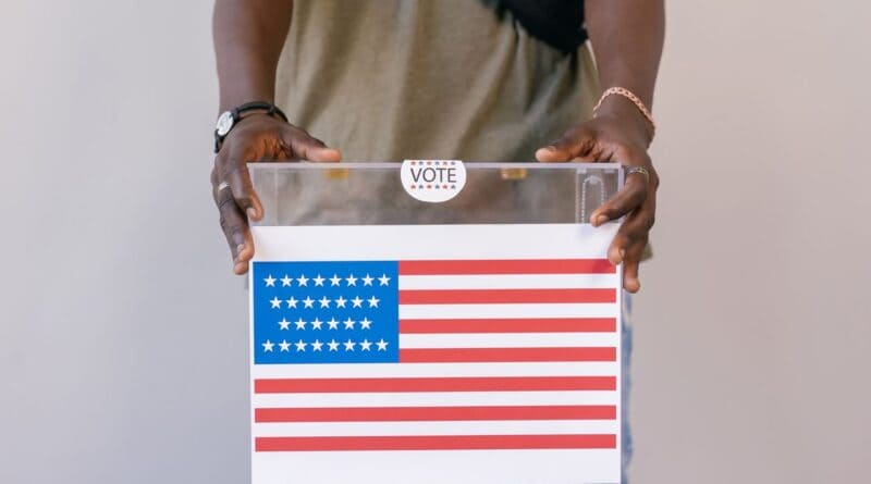 A voter places a ballot in a clear ballot box, illustrating the Corinth mayoral race