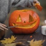 The 5 Best Pumpkin Carving Kits for Alcorn County Families A person carving a pumpkin into a jack-o'-lantern on an outdoor wooden table with autumn leaves.